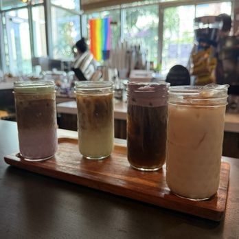 Coffee Sarap - Drink Flight - Four drinks with varying colors and textures are arranged on a wooden tray inside a cafe. The drinks are served in small glass jars, each with a different appearance, possibly indicating different flavors or ingredients. The background shows a bright, airy interior with large windows, a barista, and a rainbow pride flag, suggesting a welcoming and diverse environment. - Coffee & Tea Near Me - Denver, Colorado