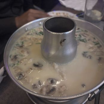 Thai Lada - Tom Kha Soup Large (With Rice) - A large metal pot filled with Tom Kha soup, a Thai coconut milk-based dish, is placed on a dining table. The soup appears creamy with visible mushrooms and herbs. The pot has a central chimney for serving. In the background, part of a person's hand and other table items, such as glasses and plates, are visible, indicating a restaurant setting. - Thai Near Me - Austin, Texas
