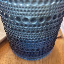 This is the most amazing cup I have ever drank from. That feels weird to say but still - The image shows a close-up of a blue glass tumbler with a textured surface featuring numerous raised bubbles or dots. The tumbler is positioned on a wooden table, and part of a white object is visible on the left side of the image. The focus is on the detailed surface of the glass, highlighting its pattern and transparency.
