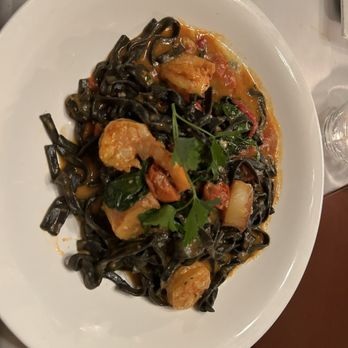 Ristorante Euno - Squid ink pasta - A white oval plate features squid ink pasta mixed with seafood, including shrimp and possibly scallops, topped with fresh herbs such as parsley. The dish is served with a tomato-based sauce and garnished with a sprig of parsley, placed on a table with a fork and glass nearby. - Italian Near Me - Boston, Massachusetts