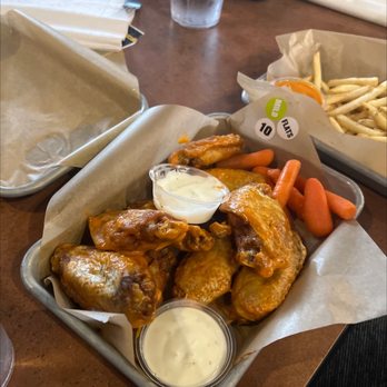 Buffalo Wild Wings - Wings - A metal basket lined with parchment paper contains buffalo chicken wings coated in sauce, accompanied by two small cups of creamy dipping sauces. Next to the basket, there are fresh carrot sticks. In the background, another basket of French fries with a side of dipping sauce is visible. The setting appears to be a casual dining restaurant with a wooden table surface. - Sports Bars Near Me - Springfield, Oregon
