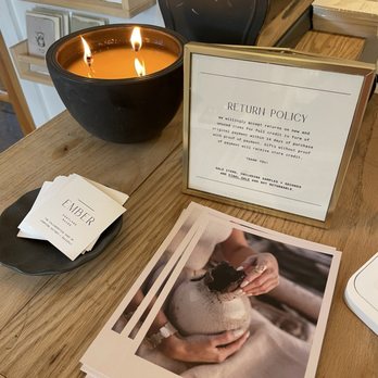 Ember - A lit candle in a black bowl, emitting a warm glow, is placed on a wooden counter. Next to it, a framed sign displays Ember's return policy, and a small black dish holds business cards with Ember's branding. Several brochures featuring a person holding a ceramic object are stacked on the counter. The background includes a display with greeting cards and a cash register, indicating a retail environment focused on handmade ceramics and artisan products. - Jewelry Near Me - Portland, Maine