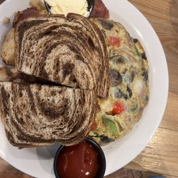 Eli's - Truffle omelet - A breakfast plate includes two slices of marble rye toast, an omelette with visible vegetables such as tomatoes and green peppers, mashed potatoes, a small container of butter, and a small container of ketchup. The food is arranged on a white plate placed on a wooden table. - Breakfast & Brunch Near Me - Natick, Massachusetts