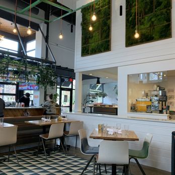Everly - Indoor seating - A cafe interior featuring high ceilings, hanging light bulbs, a large window, and a visible kitchen area with staff preparing orders. - New American Near Me - Madison, Wisconsin
