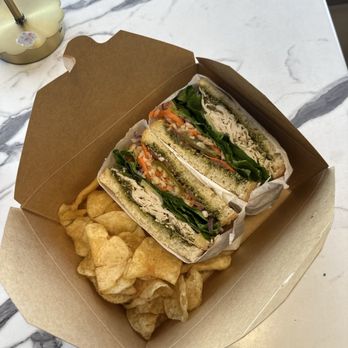 Kahlo Cafe - Turkey sandwich with pesto - A brown takeout box holds a veggie sandwich with lettuce, tomato, and other vegetables, along with potato chips, on a marble surface. - Bakeries Near Me - Oceanside, California