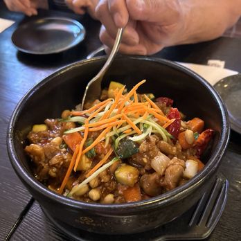 Dumpling Cafe - Kung Pio Chicken Bibimbap and shrimp dumplings - A black bowl contains stir-fried vegetables and chicken, topped with shredded carrots and other vegetables, served at a restaurant. - Dim Sum Near Me - Santa Fe, New Mexico