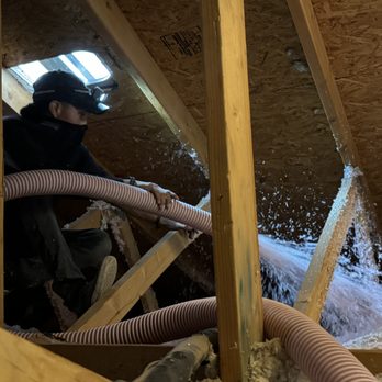 Cool Spaces Insulation - The second guy crawled all over the the attic space blowing it several inches thick. - A person wearing a headlamp and dark clothing is operating a hose to blow insulation into an attic space. The attic has exposed wooden framing, a skylight providing natural light, and insulation material being distributed through the hose. The worker is positioned on the floor of the attic, focused on the task of insulating the ceiling area. - Insulation Installation Near Me