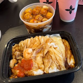 Fu Yuan - Shrimp rice roll and rice rolls with fish balls and peanut sauce - The image shows a takeout meal with a black plastic container holding rice rolls with seafood, topped with sauce and a small amount of tomato. Behind it, there is a clear plastic cup filled with fish balls in a brown sauce, sprinkled with sesame seeds. Two pink cups with black floral designs are also visible, likely containing beverages. The food is placed on a dark surface, suggesting a quick meal from a Chinese restaurant specializing in rice rolls and fish balls. - Chinese Near Me - Flushing, New York