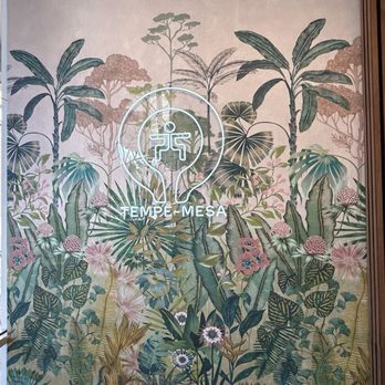 The Foundry - The image shows a decorative wall mural with detailed illustrations of tropical plants, including large leaves, flowers, and trees. A neon sign with a logo and the text 'TEMPE-MESA' is centered on the mural, illuminating the scene. The mural has a botanical theme with a variety of greenery and floral elements, creating a lush, jungle-like atmosphere. - Yoga Near Me - Mesa, Arizona