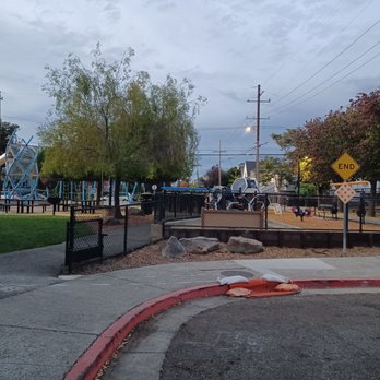 Grove Park - A playground area is enclosed by a black metal fence, featuring a slide, climbing structure, and swings. There are trees with green and reddish leaves surrounding the playground, and residential houses are visible beyond the trees. The sky is overcast, and the ground near the sidewalk has some construction sandbags and a curb painted red. - Parks Near Me - Berkeley, California