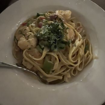M Restaurant - A bowl of seafood pasta featuring linguine noodles, shrimp, and vegetables in a creamy sauce. The dish is garnished with fresh herbs and served in a white, wide-rimmed bowl. A spoon rests on the edge of the bowl, and a glass of water is visible in the background, indicating a dining setting. - Italian Near Me - Avalon, California