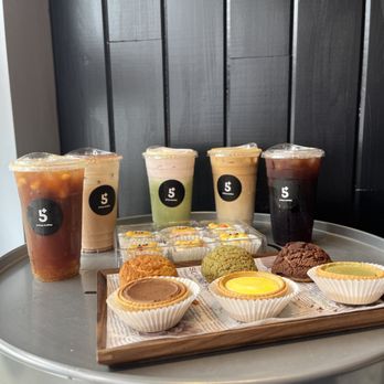 5Plus Coffee - A tray holds four iced beverages and an assortment of pastries, including egg tarts and cookies, on a metallic table against a dark wooden wall. - Coffee & Tea Near Me - Markham, Ontario