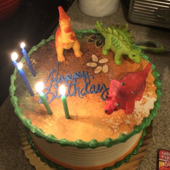 Ganache Bakery - A round birthday cake with green and orange icing features dinosaur toys and four lit candles, with a 'Happy Birthday' message. - Bakeries Near Me - Morton Grove, Illinois