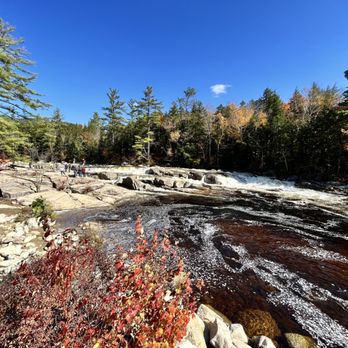 Lower Falls - A scenic outdoor scene features a river with flowing water, surrounded by rocks and a dense forest of trees. The sky is clear and blue, indicating a sunny day. Some people are visible in the distance near the riverbank, enjoying the natural setting. The trees display a mix of green and autumnal colors, suggesting a fall season. - Parks Near Me - Conway, New Hampshire