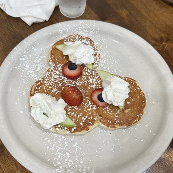 Breakfast House - The silly pancake. - Three pancakes are arranged on a white ceramic plate, each topped with whipped cream, sliced strawberries, blueberries, and small pieces of melon. The pancakes are dusted with powdered sugar. The plate is on a wooden table, with a glass of water, a napkin, and condiments visible in the background. - Breakfast & Brunch Near Me - Chicago, Illinois