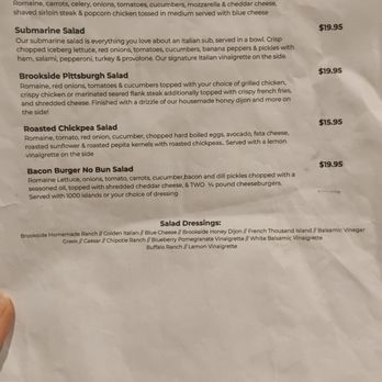 The Brookside - Salad specials menu December 2025 - A paper menu titled 'Salad Specials' lists five salads: Stinger Salad, Submarine Salad, Brookside Pittsburgh Salad, Roasted Chickpea Salad, and Bacon Burger No Bun Salad. Each item includes a detailed description of ingredients and prices ranging from $15.95 to $19.95. The menu also features a section for salad dressings, listing options such as Ranch, Italian, Blue Cheese, and others. The menu appears slightly crumpled and is held at the bottom left corner. - American Near Me - Newfane, New York