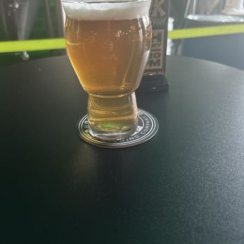 Ghost Hawk Brewing - A clear glass of beer with a foamy head sits on a black table, with brewery equipment visible in the background. - Breweries Near Me - Clifton, New Jersey