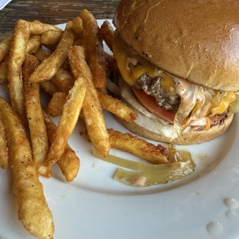The Joint - The Joint Burger - A cheeseburger with a toasted bun, lettuce, tomato, melted cheese, and a beef patty is served on a white plate. French fries are placed next to the burger, with some fries overlapping the plate's edge. The background shows a wooden table and a plate with additional food items. The image captures a typical American-style meal in a casual dining setting. - Beer, Wine & Spirits Near Me - Boston, Massachusetts