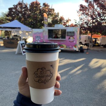 Sugar N Glitz - Hot chocolate - A person holds a coffee cup with a cardboard sleeve in front of a pink food truck at an outdoor market during daylight. - Bakeries Near Me