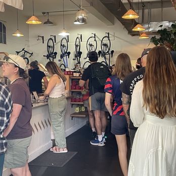 The Nitro Bar- Providence - The image shows a group of people standing in line inside a coffee shop, which features bicycle-themed wall art and hanging pendant lights. The interior has a modern, industrial style with exposed ceiling elements and a mix of casual and trendy clothing among the customers. Some individuals are engaged with the counter, while others wait patiently, indicating a busy environment. - Coffee & Tea Near Me - Providence, Rhode Island