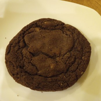 Planet Coffee - Chocolate crunch cookie - A single chocolate crunch cookie with a rough, cracked surface and a dusting of cocoa powder is positioned on a white plate. The cookie appears thick and textured, with visible cracks and uneven edges, set on a wooden surface. - Desserts Near Me - Ottawa, Ontario