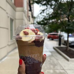 Acai Bowl 12 Oz - A hand with red-painted nails holds a large acai bowl from Deep Purple, topped with whipped cream and berries, on a city sidewalk with trees and buildings in the background.