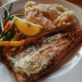 Tangled Garden 2.0 - Honey dijon trout - A plate contains a baked salmon fillet topped with herbs and a creamy sauce, accompanied by a serving of mashed potatoes and a side of cooked green beans with carrots. A lemon wedge is placed next to the mashed potatoes, and the dish is served on a white plate. - American Near Me - Perth, Ontario