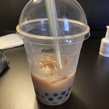 Make Waves - Brown sugar milk tea - A transparent plastic cup filled with brown sugar milk tea, containing dark tapioca pearls at the bottom and ice cubes on top, is shown on a black table. The cup has a clear plastic lid with two straws inserted, one white with green stripes and the other plain white. In the background, there is a white face mask, a small bottle, and a black surface, indicating an indoor setting. - Bubble Tea Near Me - Milwaukee, Wisconsin