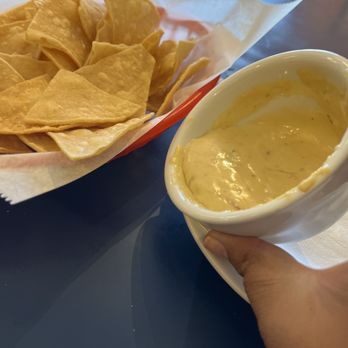 Pantry Restaurant - Chips & Queso - A basket containing tortilla chips lined with paper, placed on a dark table, with a cup of cheese dip held in a person's hand nearby. The setting appears to be a casual dining environment, likely a restaurant or cafe. - Breakfast & Brunch Near Me - Santa Fe, New Mexico