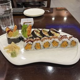 Shrimp Tempura and Sweet Potato Rollsweet - A white ceramic plate contains an assortment of sushi rolls, including tempura and mango-filled varieties, topped with sauces and garnished with lettuce. Accompanying the sushi are slices of pickled ginger and a dollop of wasabi. The plate is placed on a dark wooden table in a restaurant setting, with a glass of water, a small dipping sauce dish, and a plate and utensils visible in the background.