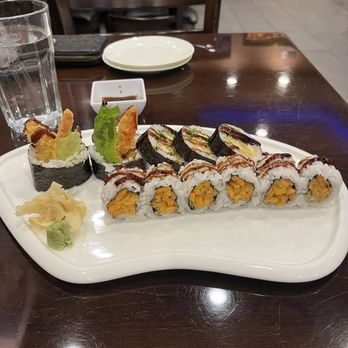Sakura Grill - Shrimp Tempura and Sweet Potato Rollsweet - A white ceramic plate contains an assortment of sushi rolls, including tempura and mango-filled varieties, topped with sauces and garnished with lettuce. Accompanying the sushi are slices of pickled ginger and a dollop of wasabi. The plate is placed on a dark wooden table in a restaurant setting, with a glass of water, a small dipping sauce dish, and a plate and utensils visible in the background. - Sushi Bars Near Me - Burke, Virginia