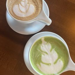 Matcha & vanilla latte - Two white cups of coffee beverages, one with latte art and the other with matcha latte art, are placed on a wooden table.