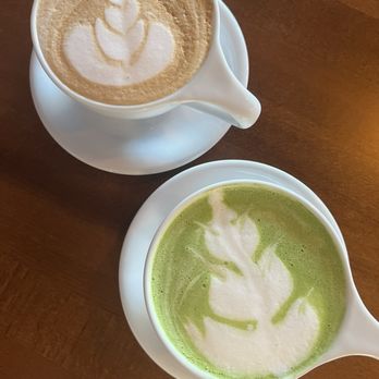 Dancin’ Beans Cafe - Matcha & vanilla latte - Two white cups of coffee beverages, one with latte art and the other with matcha latte art, are placed on a wooden table. - Coffee & Tea Near Me - Euless, Texas