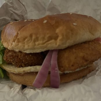 Mad Chicken - A fried chicken sandwich with pickled onions, lettuce, and sauce on a bun, wrapped in paper, likely from a casual eatery. - Chicken Wings Near Me - Milwaukee, Wisconsin