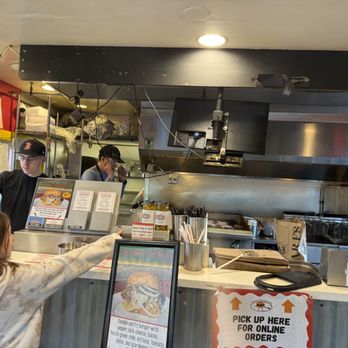 Snarfburger - the counter and grill - The image shows a restaurant counter with two staff members behind it, one wearing a black cap and the other with glasses, working in the kitchen area. A young girl with a hair clip is pointing at the menu or counter, and a person is taking a photo or video with a phone. The counter has signs, utensils, and containers, with a sign indicating online order pickup. The kitchen area includes cooking equipment and shelves with supplies. - Burgers Near Me - Boulder, Colorado