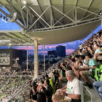 Q2 Stadium - A large crowd of spectators is seated in a stadium with green seats, watching an event on the field below. The stadium has a modern roof structure with visible metal beams and lighting. The background shows a city skyline with tall buildings and a colorful sunset sky with pink and purple hues. - Stadiums & Arenas Near Me - Austin, Texas