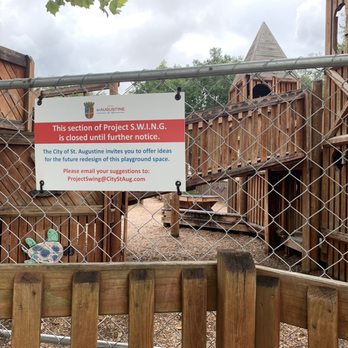 Project SWING Park - Did this really *need* to be redesigned? - The image shows a fenced playground area made of wooden structures, including a small tower and stairs. A sign attached to the fence indicates that this section of Project S.W.I.N.G. is closed until further notice. The sign invites suggestions for future redesigns and provides an email address for submissions. The background includes cloudy skies and trees, with the playground equipment and fencing occupying most of the scene. - Parks Near Me - St. Augustine, Florida