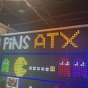 Pins Mechanical - A LED sign with the text 'FINS ATX' in white and yellow lights, featuring symbols like a dollar sign, a fish, and other designs, set against a dark ceiling. - Bowling Near Me - Austin, Texas