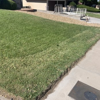 Empire Yard Maintenance - The image shows a patch of well-maintained grass next to a concrete sidewalk. The grass has a slight slope and is bordered by a small strip of dirt. In the background, there is a building with a ramp, some landscaping with bushes, and a gravel area. The scene appears to be part of a residential or commercial property with outdoor landscaping. - Landscaping Near Me