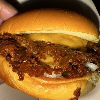 Twans Burger - A close-up of a burger with crispy bacon, melted cheese, and white onions inside a soft, toasted bun. The burger appears freshly prepared and is held in a person's hand, with a background suggesting a casual dining environment. - Food Trucks Near Me - Denver, Colorado