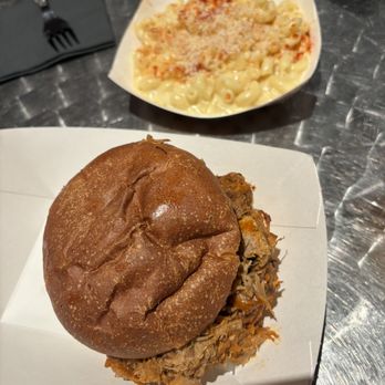 Cafe Berlin - Pulled pork sandwich, Mac n cheese - A pulled pork sandwich with a round, toasted bun is served on a white paper tray, with shredded pork visible underneath. In the background, a bowl of macaroni and cheese topped with cheese and breadcrumbs is placed on a metal table. The setting appears to be a casual dining environment with utensils and a black napkin nearby. - Breakfast & Brunch Near Me - Columbia, Missouri