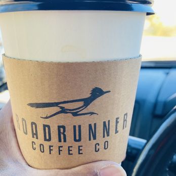 Roadrunner Coffee - Vanilla Gingerbread Latte - A disposable coffee cup with a black plastic lid is held in a person's hand inside a vehicle. The cup has a cardboard sleeve with the Roadrunner Coffee Co. logo, which includes a stylized roadrunner bird. The background shows part of the vehicle's interior and a blurred outdoor scene. - Coffee & Tea Near Me - Tucson, Arizona