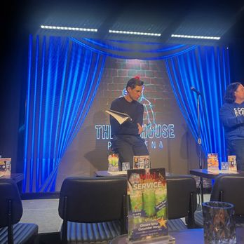 Cool Beans Comedy - Two individuals are on stage at The Ice House Comedy Club in Pasadena, with a large screen displaying the venue's name behind them. The stage has blue curtains and promotional materials on tables. - Performing Arts Near Me - Pasadena, California
