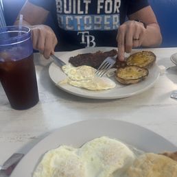 Corned beef hash, biscuit eggs - A person wearing a dark T-shirt with the text 'BUILT FOR OCTOBER' is seated at a white table, using a fork and knife to eat breakfast. The plate contains fried eggs, roasted potatoes, and a meat dish. A bowl of mashed potatoes or rice is visible to the right. A glass of dark soda with a straw is on the left side of the table. The setting appears to be a casual restaurant or diner.