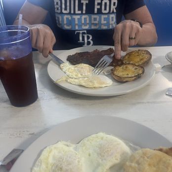 Apollo Beach Diner - Corned beef hash, biscuit eggs - A person wearing a dark T-shirt with the text 'BUILT FOR OCTOBER' is seated at a white table, using a fork and knife to eat breakfast. The plate contains fried eggs, roasted potatoes, and a meat dish. A bowl of mashed potatoes or rice is visible to the right. A glass of dark soda with a straw is on the left side of the table. The setting appears to be a casual restaurant or diner. - Diners Near Me - Apollo Beach, Florida