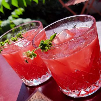 OTB - Cherry bomb - Two pink cocktails with ice cubes and small green garnishes are positioned on a reddish-brown table outdoors. The glasses have a textured, geometric pattern, and condensation is visible on the surface. The background features green foliage, indicating an outdoor setting, possibly at a bar or restaurant patio. - New American Near Me - Brooklyn, New York