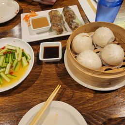 Summer Rolls - A variety of Asian dishes are displayed on a wooden table, including steamed buns, spring rolls with dipping sauces, and a cucumber salad.