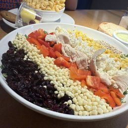 Chopped Salad - A large salad on a white oval plate contains shredded chicken, chopped tomatoes, corn, dried cranberries, and cheese. The salad is arranged with each ingredient in its own section. In the background, there is a bowl of macaroni salad, a slice of bread, and a small dish of dressing on a wooden table. A person wearing a blue shirt is partially visible in the background.
