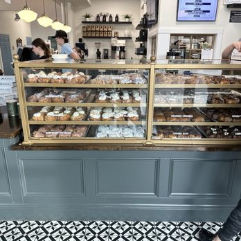 Parlor Doughnuts - Picture of the lovely doughnuts. If you zoom in you can see the doughnuts and the names posted. - A glass doughnut display case with a gold frame contains multiple trays of assorted doughnuts, each labeled with flavor names such as 'Strawberry', 'Blueberry Hill', and 'Chocolate Chocolate'. The case is positioned on a blue-gray counter with decorative paneling, and the background shows staff behind the counter and customers waiting. The setting appears to be inside Parlor Doughnuts in St. Augustine, FL, with a patterned tile floor and a modern interior design. - Donuts Near Me - St. Augustine, Florida