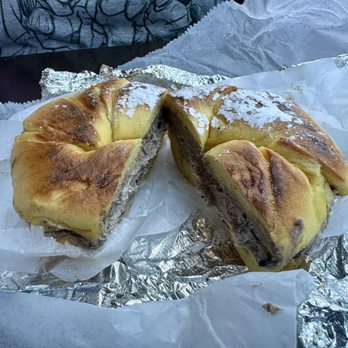 Utopia Bagels - "French Toast" bagel with Oreo cream cheese - A bagel with a chocolate filling is shown, cut in half to reveal the dark chocolate spread inside. The bagel has a golden-brown crust and is dusted with powdered sugar on top. It rests on crinkled aluminum foil and white paper, with a blurred background including a person wearing a patterned shirt. The setting suggests a casual dining or takeout environment at a bagel shop. - Bakeries Near Me - Whitestone, New York